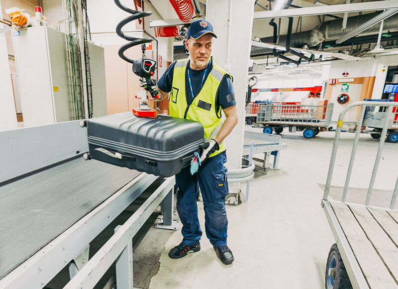 Man som lyfter resväska från transportband med hjälp av ett lyfthjälpmedel.